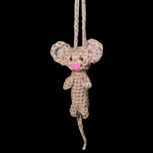 May include: A handmade crochet mouse ornament with a pink nose and large ears. The mouse is light brown and gray, with a long tail and a loop for hanging. The background is black.