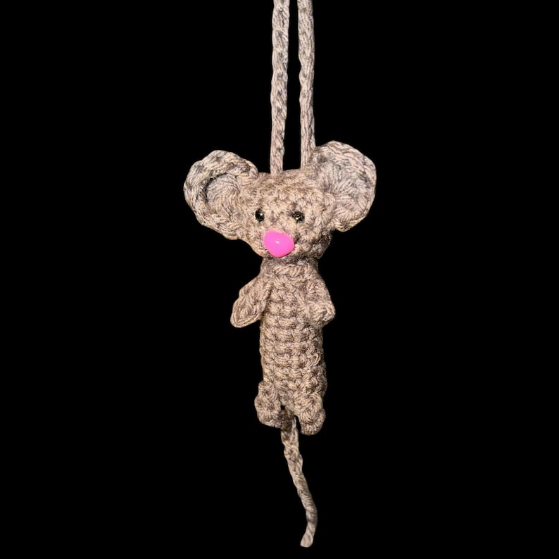 May include: A handmade crochet mouse ornament with a pink nose and large ears. The mouse is light brown and gray, with a long tail and a loop for hanging. The background is black.