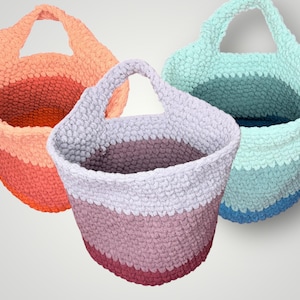 May include: Three handmade crochet baskets in shades of orange, pink, and blue. Each basket has a rounded shape with a handle and a striped color pattern. The baskets are made of a thick, textured yarn.