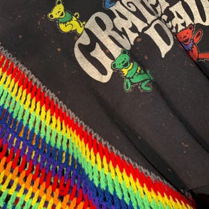 Op de afbeelding: Zwart T-shirt met de woorden "Grateful Dead" in verweerde witte letters. Kleurrijke dansende beren zijn over het shirt verspreid. Een regenboogkleurige, gehaakte sjaal is zichtbaar in de linker benedenhoek, met een grijze rand.