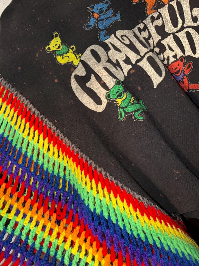 Op de afbeelding: Zwart T-shirt met de woorden "Grateful Dead" in verweerde witte letters. Kleurrijke dansende beren zijn over het shirt verspreid. Een regenboogkleurige, gehaakte sjaal is zichtbaar in de linker benedenhoek, met een grijze rand.