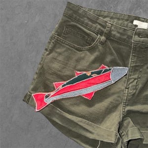 May include: Olive green denim shorts featuring a large, stitched fabric patch in the shape of a fish. The fish patch is constructed from layered red, dark green, and grey fabric pieces with visible white stitching along the edges of the design.