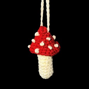 May include: A handmade crochet mushroom ornament. The mushroom cap is red with white spots, and the stem is cream-colored. A cream-colored string is attached for hanging. The background is black.