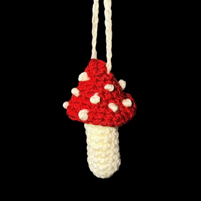 May include: A handmade crochet mushroom ornament. The mushroom cap is red with white spots, and the stem is cream-colored. A cream-colored string is attached for hanging. The background is black.