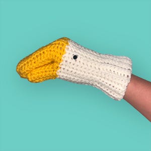 May include: A white and yellow crocheted hand puppet shaped like a goose. The puppet has a black eye and a yellow beak.