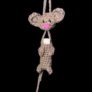 May include: A handmade gray crochet mouse holding a white object, possibly a lip balm or small container. The mouse has a pink nose and large ears. The background is solid black.