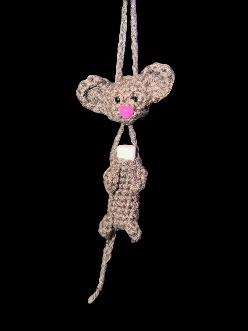 May include: A handmade gray crochet mouse holding a white object, possibly a lip balm or small container. The mouse has a pink nose and large ears. The background is solid black.