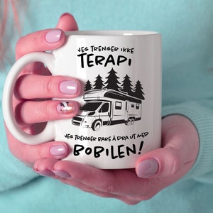 以下が含まれることがあります： 白いセラミックマグカップ。黒いキャンピングカーのグラフィックとノルウェー語のテキストが描かれています。テキストは「セラピーは必要ない、キャンピングカーで出かけるだけ！」と書かれています。ピンクのマニキュアをした人がマグカップを持っています。