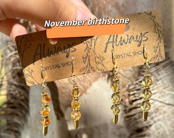May include: Three pairs of gold earrings with topaz birthstone beads. The earrings are on a brown card with the text "Always Crystal Shop" and "November birthstone".