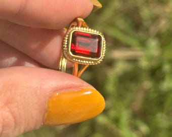 May include: A gold ring with a red gemstone set in a square bezel. The ring has a beaded edge.