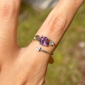 Puede incluir: Un anillo de plata con una piedra preciosa púrpura y alas de murciélago en la parte superior. El anillo está en un dedo.