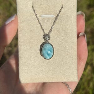 Pode incluir: Um colar de corrente de prata com um pingente oval azul que apresenta uma pedra preciosa com um padr&atilde;o de m&aacute;rmore. O pingente &eacute; engastado em uma moldura de prata com um design decorativo.