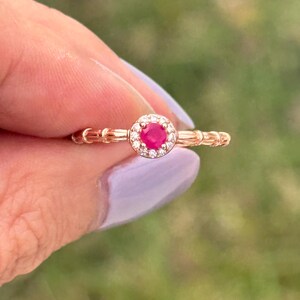 May include: A rose gold ring with a red ruby gemstone surrounded by a halo of smaller clear gemstones.