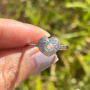 Op de afbeelding: Een zilveren ring met een hartvormige zetting met een ronde opaal-edelsteen. De ring is versierd met kleine, heldere stenen rond de opaal.