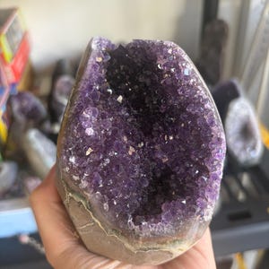 May include: A large amethyst geode featuring a deep purple, crystalline interior with jagged, multifaceted points. The exterior of the stone is a natural, rough, light brown rock formation that contrasts with the vibrant, dark violet crystal cavity.