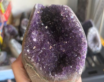 May include: A large amethyst geode featuring a deep purple, crystalline interior with jagged, multifaceted points. The exterior of the stone is a natural, rough, light brown rock formation that contrasts with the vibrant, dark violet crystal cavity.