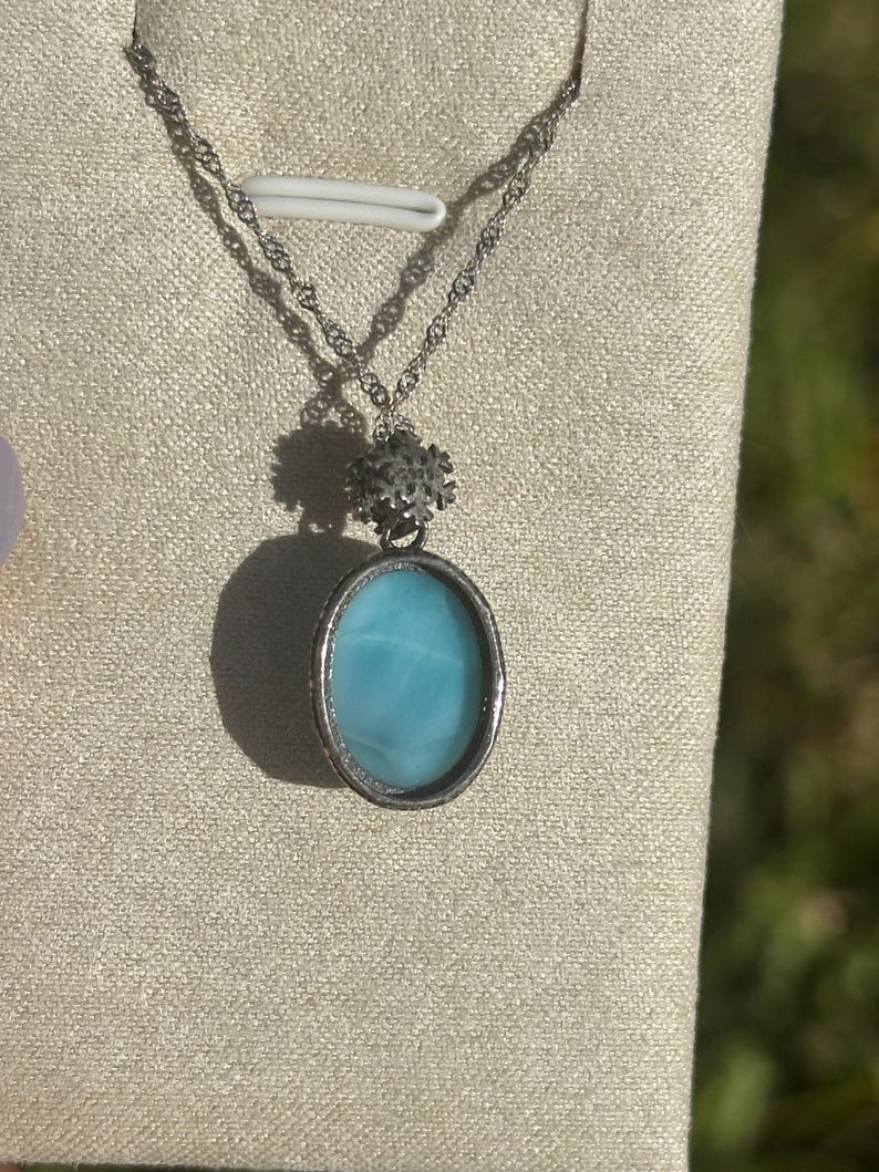 Pode incluir: Um colar de corrente de prata com um pingente oval azul e um charme de floco de neve prateado.