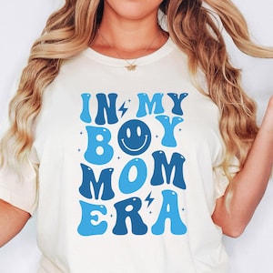 May include: A white t-shirt with blue text that reads "In My Boy Mom Era" with a blue smiley face in the middle of the text.