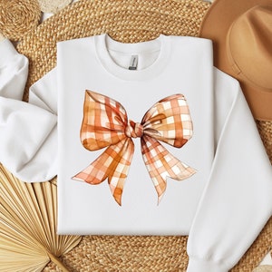 May include: White sweatshirt with a large orange and white plaid bow printed on the front.