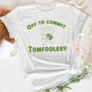 May include: White t-shirt with green text that reads "Off to commit tomfoolery". A green frog with a backpack is walking under a green leaf umbrella.