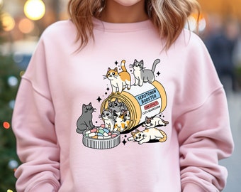 May include: Pink sweatshirt with a graphic of a pill bottle overflowing with cute cartoon cats. The pill bottle is labeled "Serotonin Booster" with the side effect listed as "Cute Aggression".