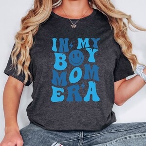 May include: A dark grey t-shirt with a blue and white graphic that says "In My Boy Mom Era" with a smiley face in the middle of the word "Boy".