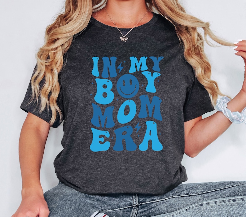 May include: A dark grey t-shirt with a blue and white graphic that says "In My Boy Mom Era" with a smiley face in the middle of the word "Boy".