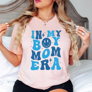May include: A pink t-shirt with the text "In My Boy Mom Era" in blue and white letters with a smiley face in the middle of the word "Boy".