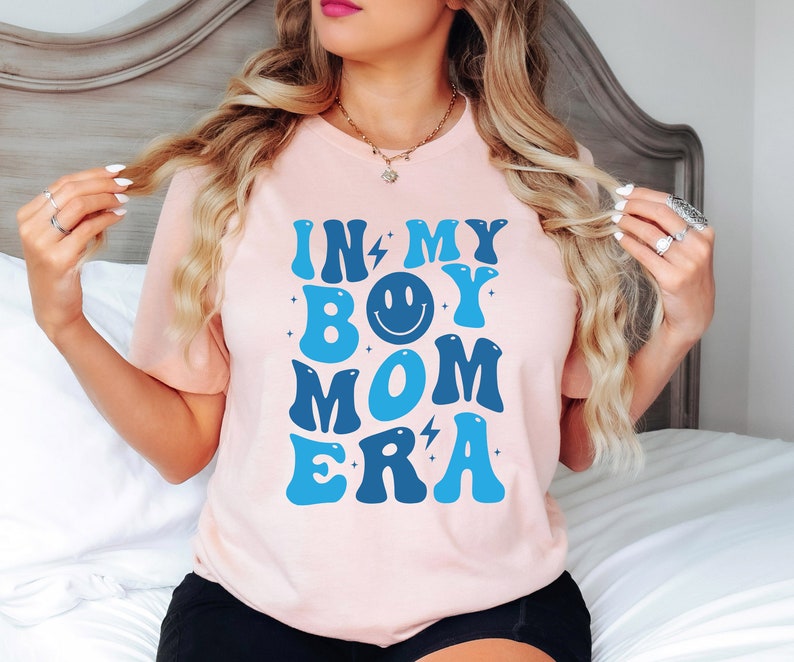 May include: A pink t-shirt with the text "In My Boy Mom Era" in blue and white letters with a smiley face in the middle of the word "Boy".