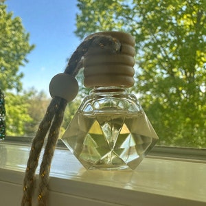 May include: A clear glass bottle with a wooden cap and a brown and gold cord. The bottle is filled with a clear liquid and has a diamond-shaped design.