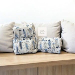 May include: Three white quilted zippered pouches with a blue and white fish pattern. The pouches are stacked on top of each other on a wooden surface.