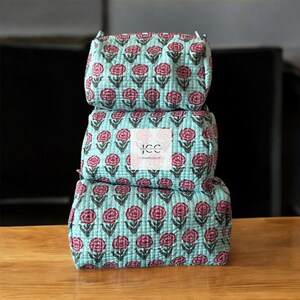 May include: A stack of three turquoise blue quilted makeup bags with a pink floral print. The bags are made of cotton and have a zipper closure. The top bag has the text "JCC Premium Quality" printed on it.