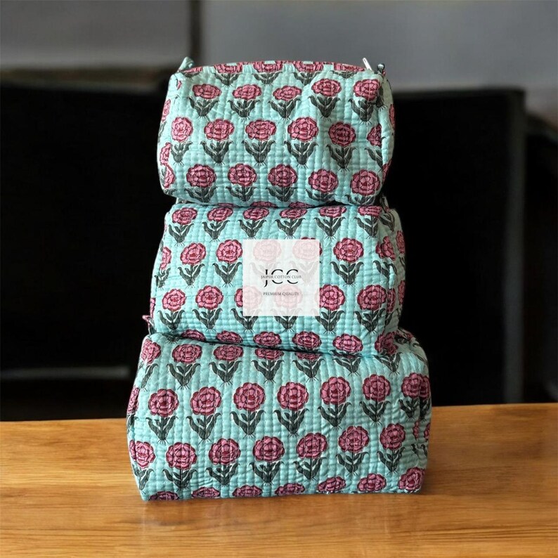 May include: A stack of three turquoise blue quilted makeup bags with a pink floral print. The bags are made of cotton and have a zipper closure. The top bag has the text "JCC Premium Quality" printed on it.