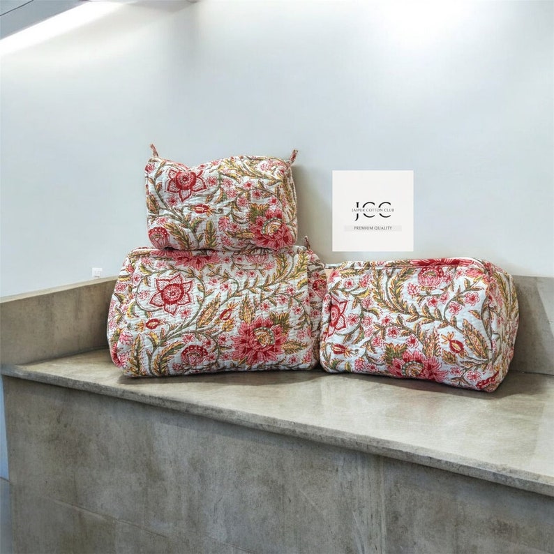 May include: Three quilted fabric cosmetic bags with a floral print in shades of pink, white, and green. The bags are stacked on top of each other on a light grey surface. The text "JCC - Jaipur Cotton Club - Premium Quality" is visible in the top right corner of the image.