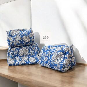 May include: Three blue and white floral print toiletry bags stacked on a wooden surface. The bags have a quilted texture and are made of cotton.
