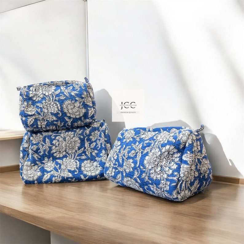 May include: Three blue and white floral print toiletry bags stacked on a wooden surface. The bags have a quilted texture and are made of cotton.