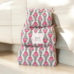 May include: Three pink and white floral print quilted makeup bags stacked on top of each other. The bags have a white background with pink flowers and grey leaves. The top bag has the text "JCC" in a white box.