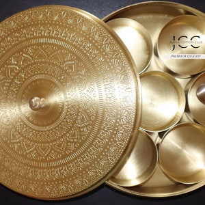 May include: A round, gold-colored metal spice box with a lid featuring an intricate design. The box has six compartments and a small spoon. The lid has the text "Jaipur Cotton Club Premium Quality".