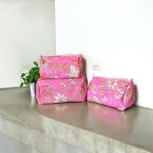 May include: Three pink quilted makeup bags with a floral print. The bags are stacked on top of each other on a light grey surface.