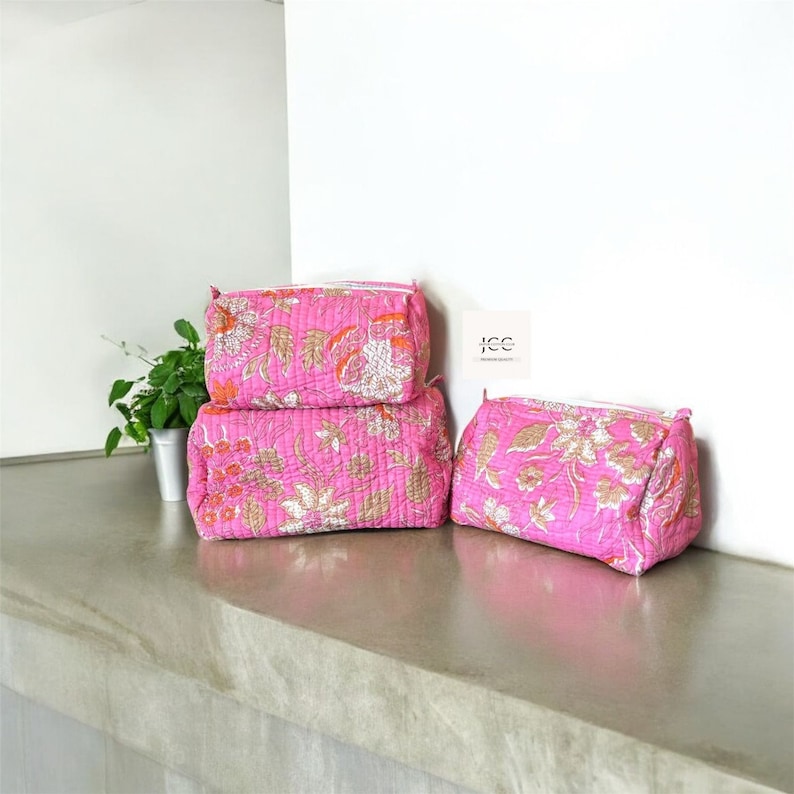 May include: Three pink quilted makeup bags with a floral print. The bags are stacked on top of each other on a light grey surface.