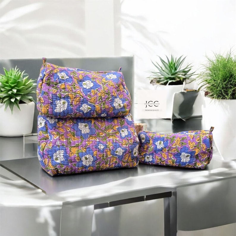May include: Three purple and white floral print fabric zippered pouches. The largest pouch is standing upright and the two smaller pouches are lying flat on a grey table.