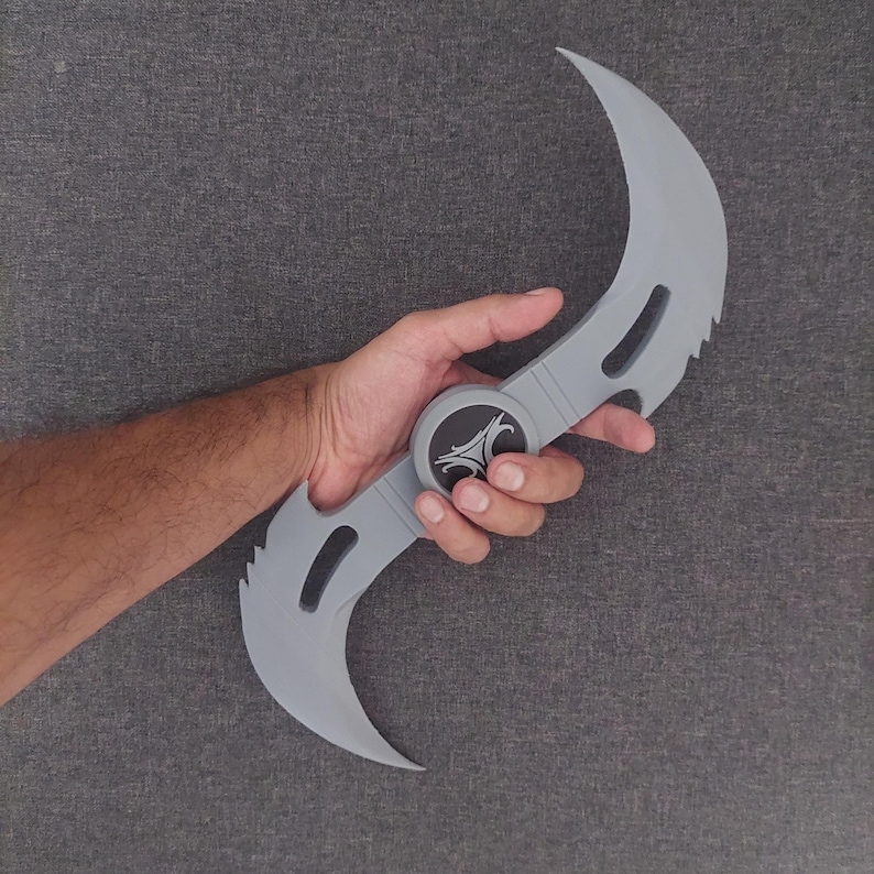 May include: A gray, double-bladed karambit knife with a circular center and a stylized emblem. The curved blades have serrated edges and a pointed tip. The knife is held in a hand against a gray background.