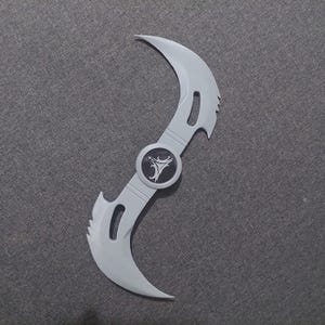 May include: A light gray, double-bladed prop weapon with a central circular emblem. The blades curve in opposite directions, each with a curved cutout. The emblem is black with a white stylized design.