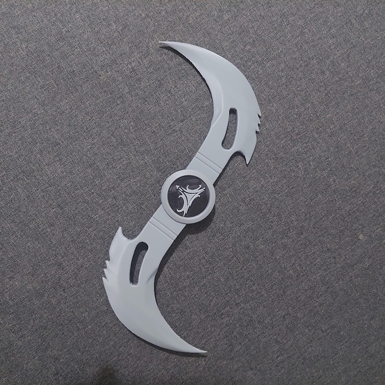 May include: A light gray, double-bladed prop weapon with a central circular emblem. The blades curve in opposite directions, each with a curved cutout. The emblem is black with a white stylized design.