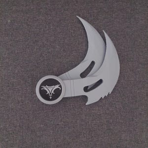 May include: A light gray karambit knife with a curved blade and a circular handle featuring a black emblem with a white design. The blade has a serrated edge. The knife is lying on a gray surface.