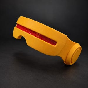 May include: A yellow visor featuring a horizontal cutout that reveals a red mesh insert. The visor has a smooth, matte finish with circular side components and a curved frame designed to contour around the face, presenting a solid structure.