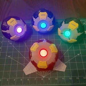 Può includere: Quattro dispositivi sferici dall'aspetto futuristico con luci centrali luminose. I dispositivi sono principalmente grigi, con accenti gialli e blu scuro. Ognuno ha una luce di colore diverso: viola, blu, verde e rosso. Sono disposti su una superficie verde.