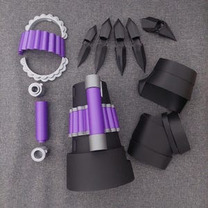 Op de afbeelding: Een verzameling zwarte en paarse cosplay-kostuumonderdelen. Inclusief zwarte vingerextensies, arm- en beenpantser en decoratieve paarse en witte accenten. De stukken zijn gerangschikt op een grijze ondergrond.
