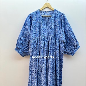 Może przedstawiać: Niebieska bawełniana sukienka z kwiatowym nadrukiem i marszczoną talią. Sukienka ma okrągły dekolt i 3/4 rękawy. Sukienka wisi na drewnianym wieszaku. Na sukience widoczny jest napis "Roeh Exports".