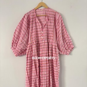 May include: A pink midi dress with a vertical striped pattern in white, black, and gold. The dress has a collared neckline, button-down front, and three-quarter sleeves. The dress is hanging on a wooden hanger.